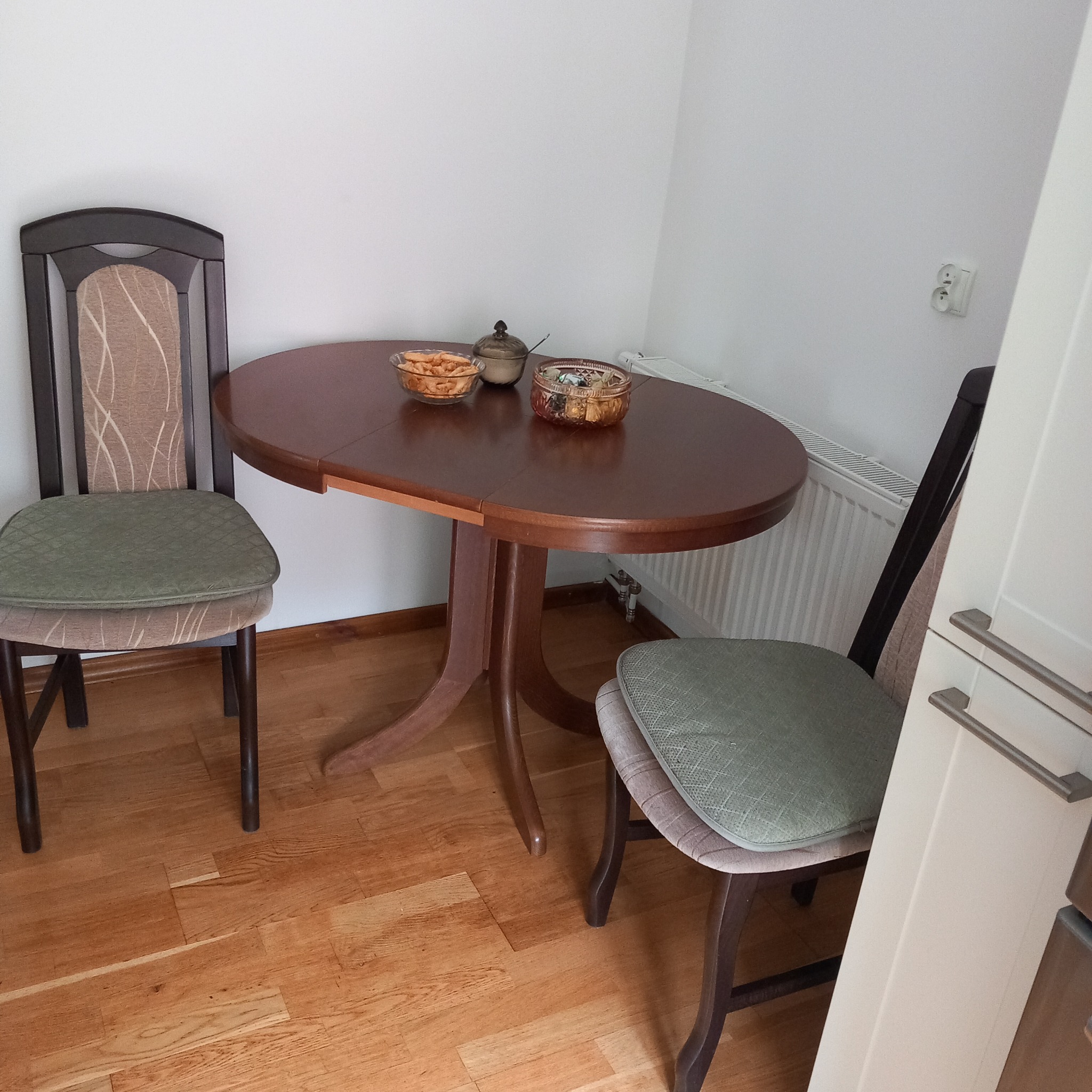 Owalny, rozkładany stół z ciemnego drewna z dwoma krzesłami o drewnianych ramach i zielonych siedziskach, na stole szklane miseczki z przekąskami i ozdobny pojemnik, parkiet na podłodze, biała...