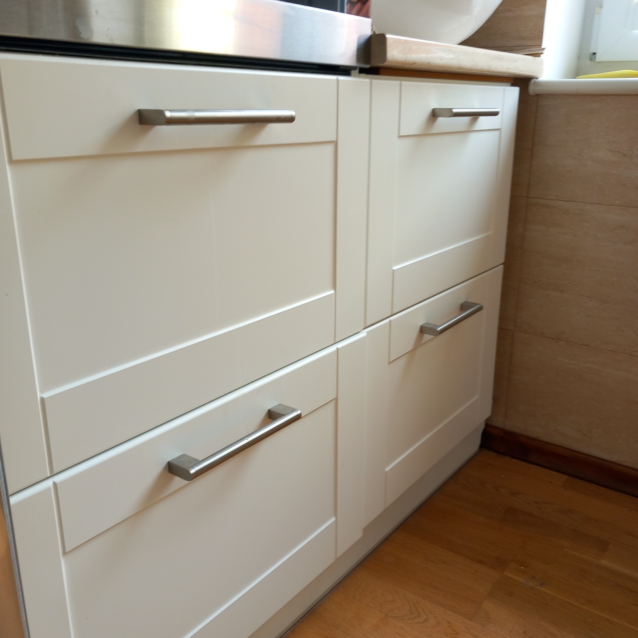 Białe, lakierowane szafki kuchenne z metalowymi uchwytami, widoczne na tle drewnianej podłogi i fragmentu ściany wyłożonej beżowymi płytkami.