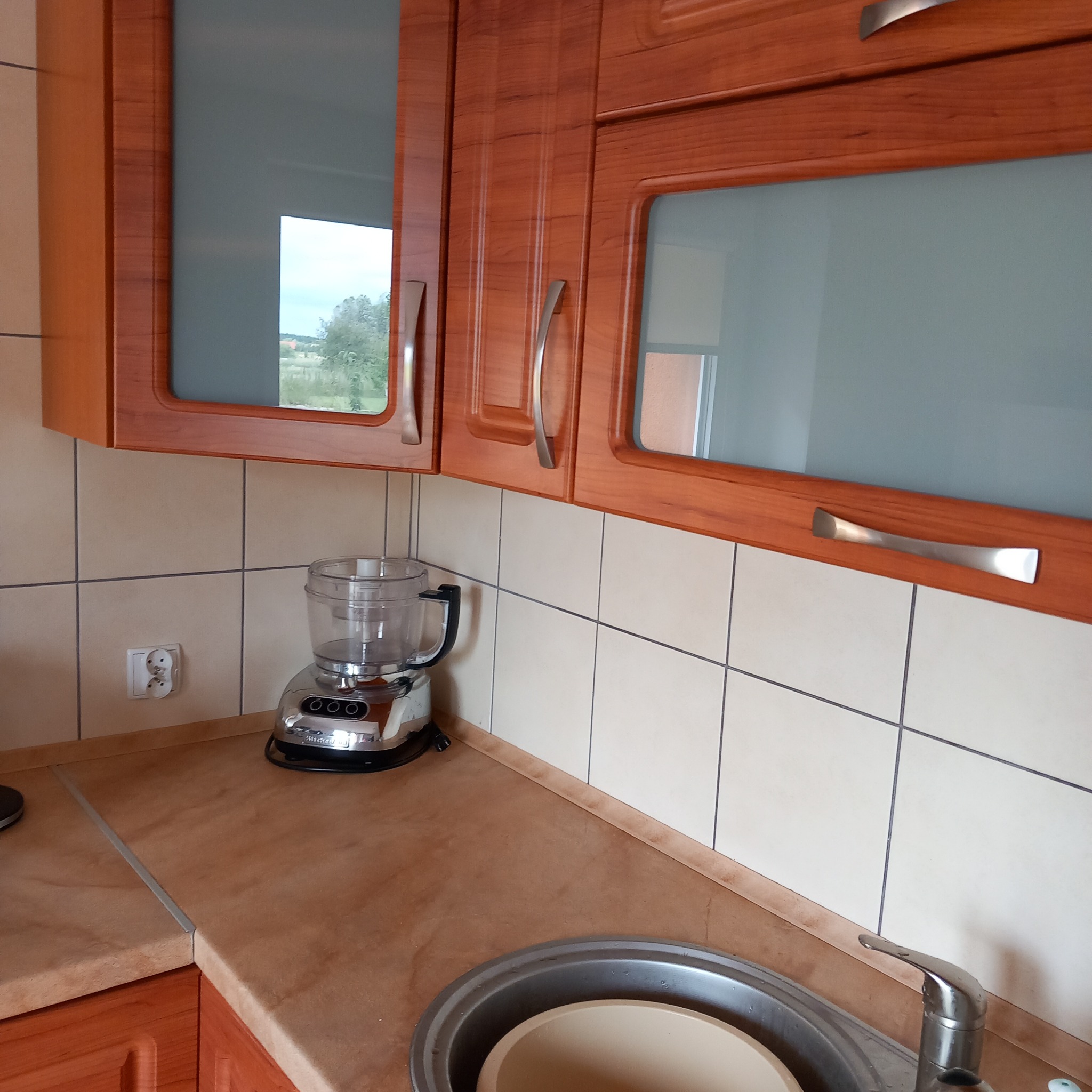 Fragment kuchni z drewnianymi szafkami wiszącymi, beżowymi kafelkami i metalowym robotem kuchennym na blacie obok zlewu.