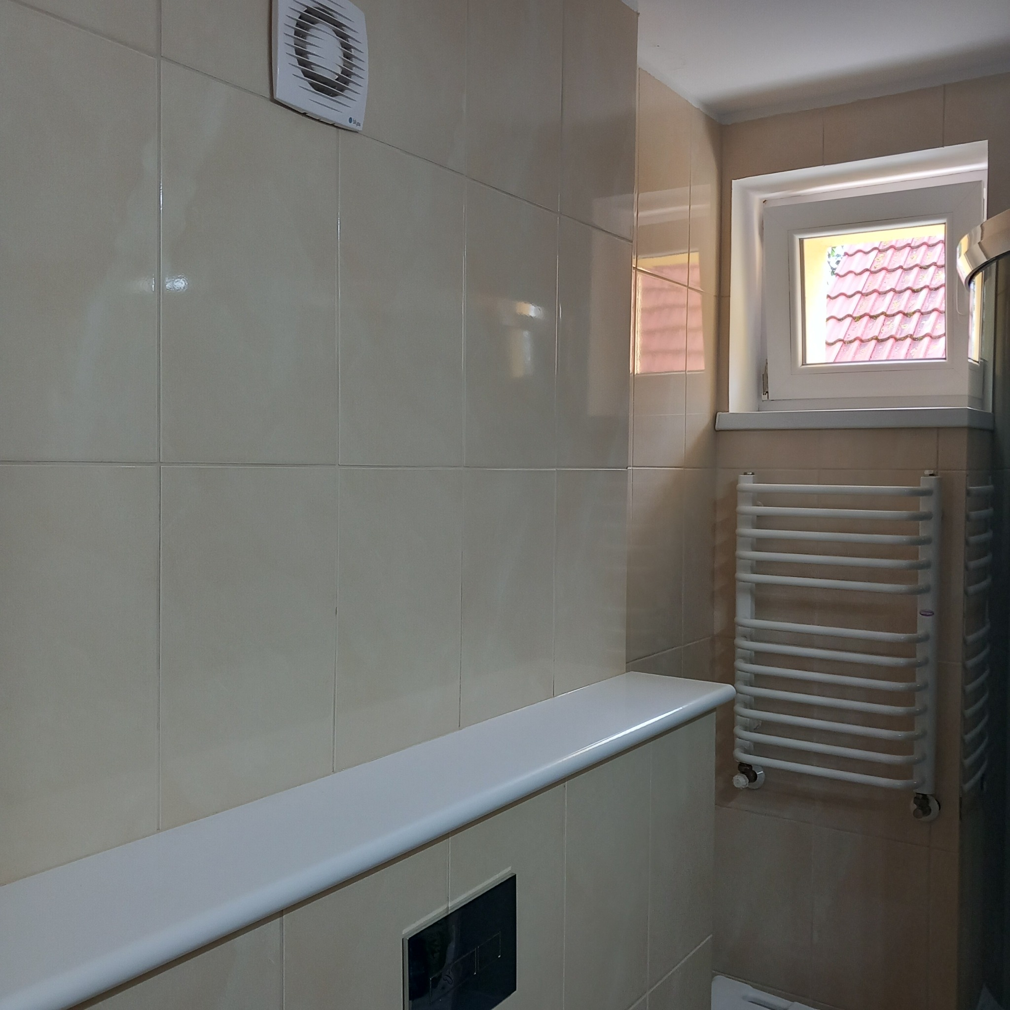 Jasna łazienka z beżowymi płytkami, białym grzejnikiem drabinkowym i oknem z widokiem na czerwony dach. Na ścianie zamontowany wentylator.