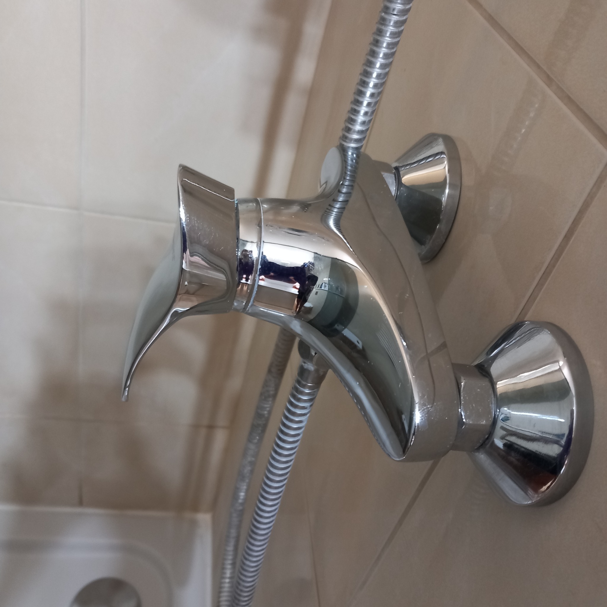 Błyszcząca, chromowana bateria prysznicowa zamontowana na beżowych płytkach ściennych w łazience, widoczny wąż prysznicowy i fragment wanny.