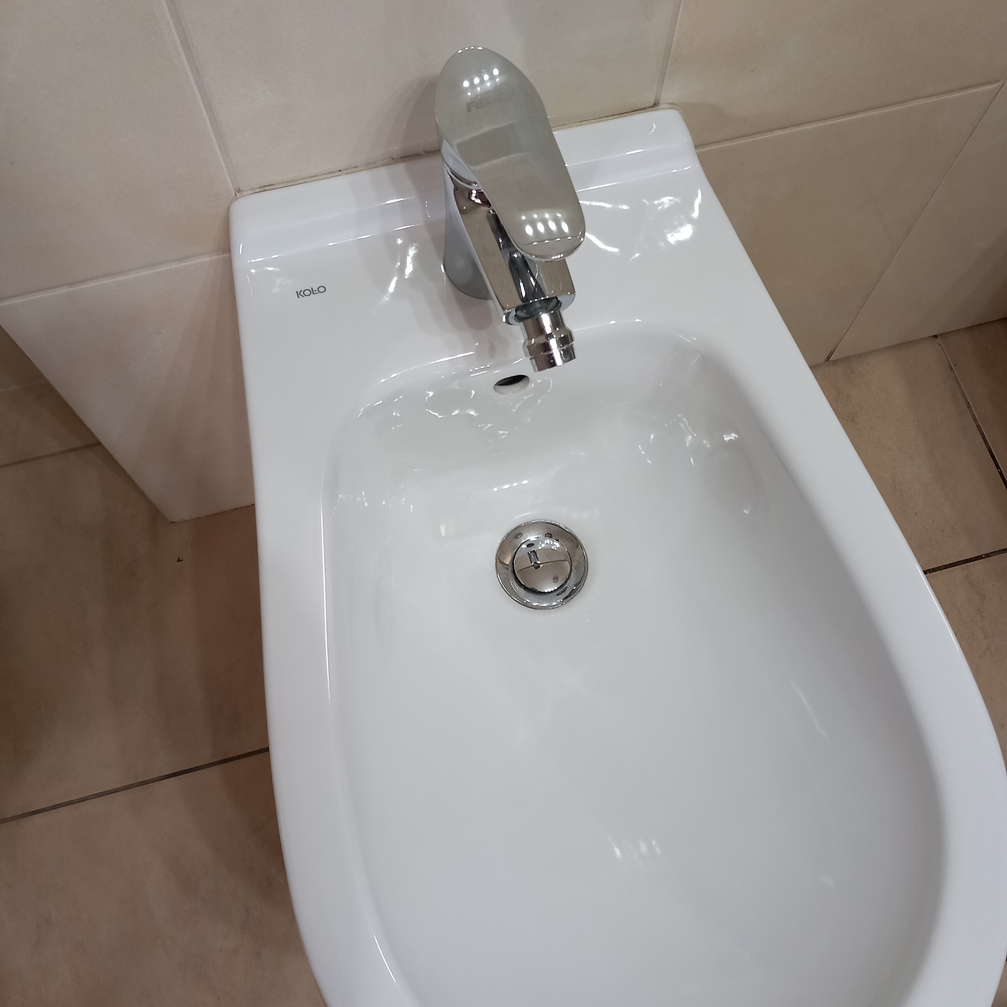 Bidet marki Koło z chromowaną baterią, zamontowany w łazience z beżowymi płytkami.