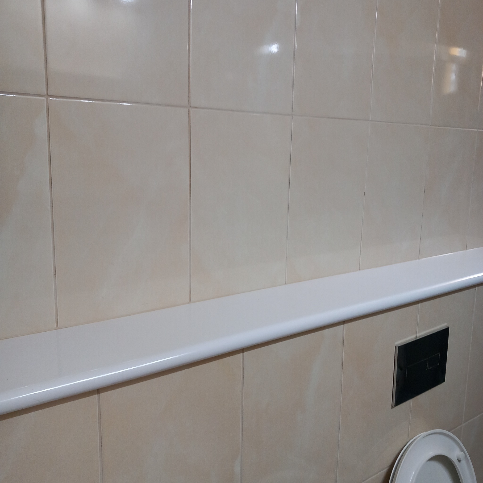 Jasne, beżowe płytki ceramiczne na ścianie łazienki, z widocznym białym obramowaniem wanny i fragmentem deski sedesowej oraz czarnym przyciskiem spłuczki.