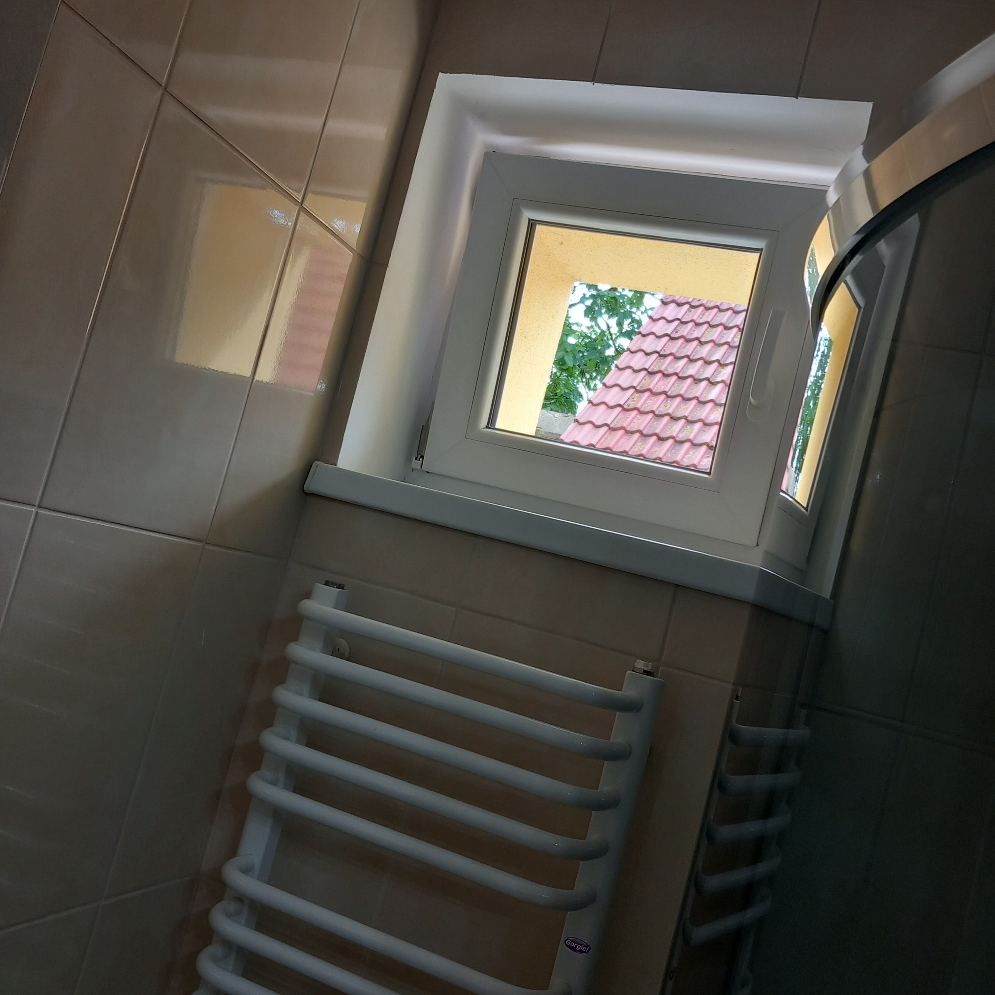 Wnętrze łazienki z widokiem na zewnątrz przez okno, na ścianach beżowe płytki, biały grzejnik drabinkowy na ścianie.