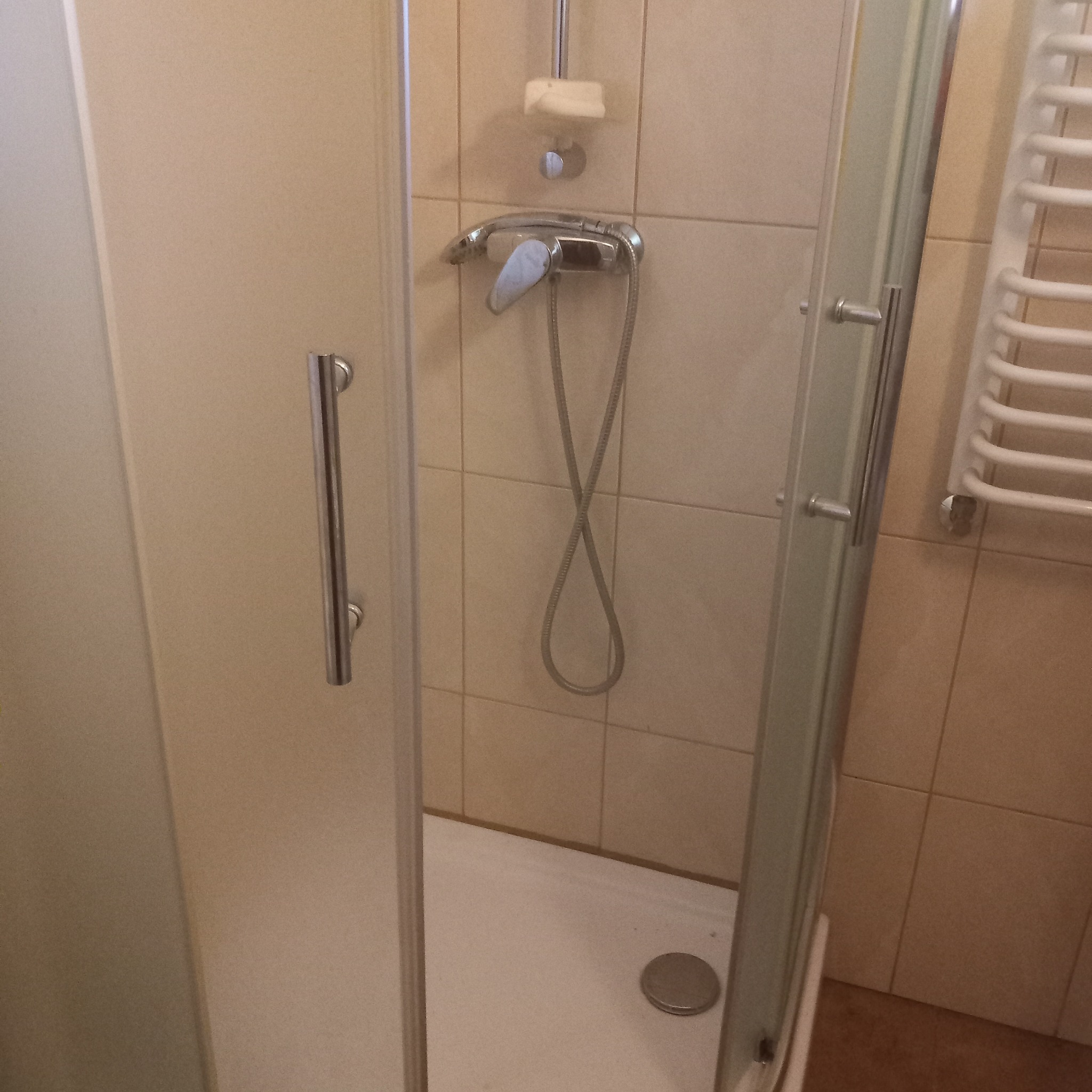 Kabina prysznicowa z chromowaną armaturą na tle beżowych płytek, widoczny odpływ i grzejnik drabinkowy.