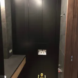 Nowoczesna łazienka z czarnymi, matowymi szafkami w zabudowie, szarą ścianą z płytek imitujących kamień, drewnianym blatem umywalkowym i ukrytą spłuczką WC z chromowanymi przyciskami.