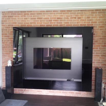 Telewizor wbudowany w czarną zabudowę meblową, wnęka wykończona cegłą, kolumny głośnikowe po bokach.