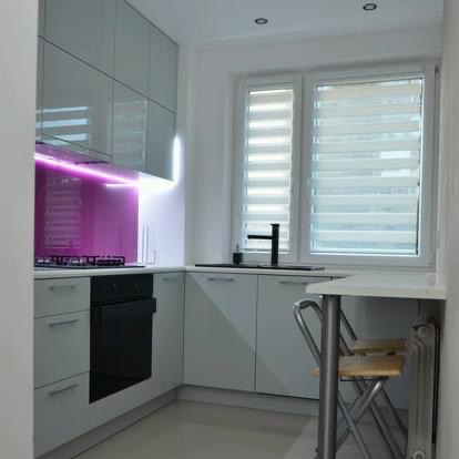 Jasnoszara kuchnia w kształcie litery L z fioletowym panelem szklanym nad płytą grzewczą, oświetlonym od góry ledowym paskiem. Widoczne okno z żaluzjami dzień-noc i czarną baterią. Dodatkowy blat...
