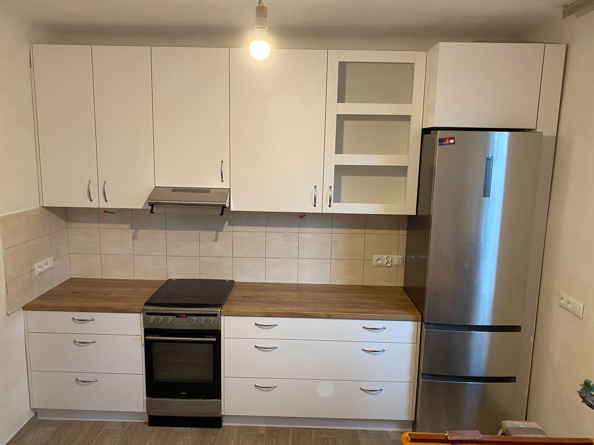 Jasna kuchnia z białymi szafkami, drewnianym blatem i srebrną lodówką, oświetlona pojedynczą żarówką