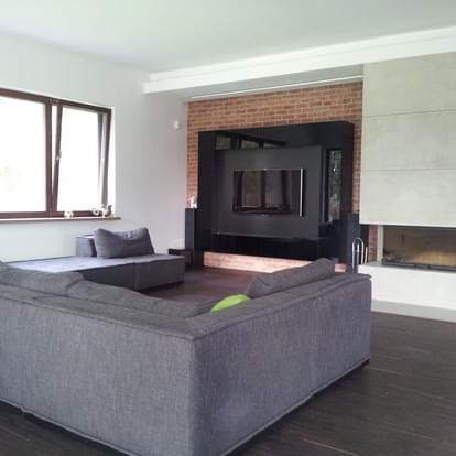 Nowoczesny salon z szarą sofą narożną, ceglaną ścianą za czarną zabudową telewizyjną z podświetleniem LED i kominkiem obok.