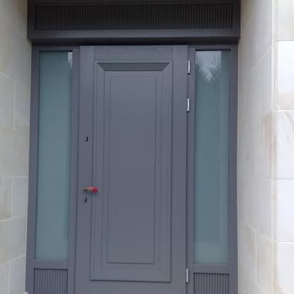 Szare drzwi wejściowe z bocznymi szybami z matowego szkła, osadzone w jasnej elewacji z kamiennych płyt.