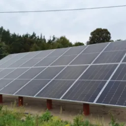 Instalacja paneli słonecznych na konstrukcji wsporczej z rur w otoczeniu zieleni i drzew, widok z boku przy pochmurnej pogodzie.