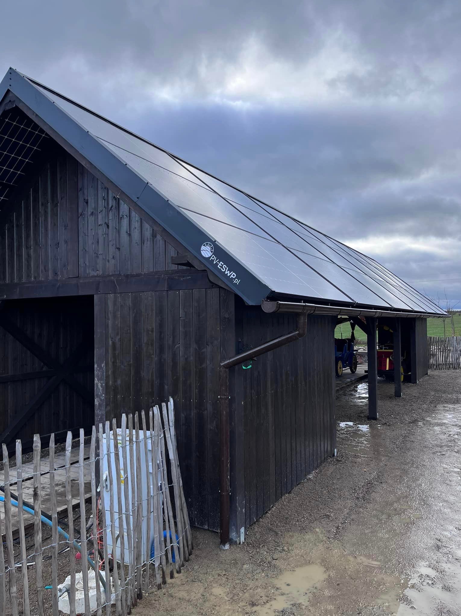 Czarna drewniana stodoła z panelami fotowoltaicznymi na dachu, widoczny z boku pod pochmurnym niebem, z logo firmy PV-ESWP.pl na panelach, teren wokół błotnisty.