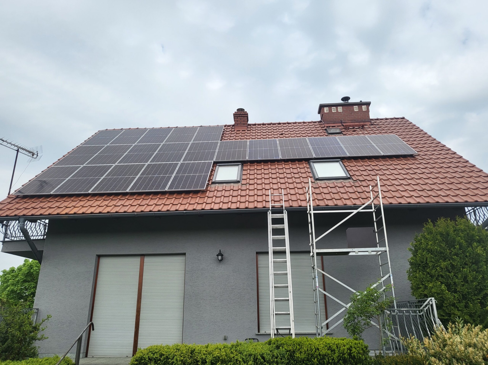 Panele słoneczne na dachu domu z czerwoną dachówką, widoczne okna dachowe i drabina oparta o elewację, pochmurne niebo.