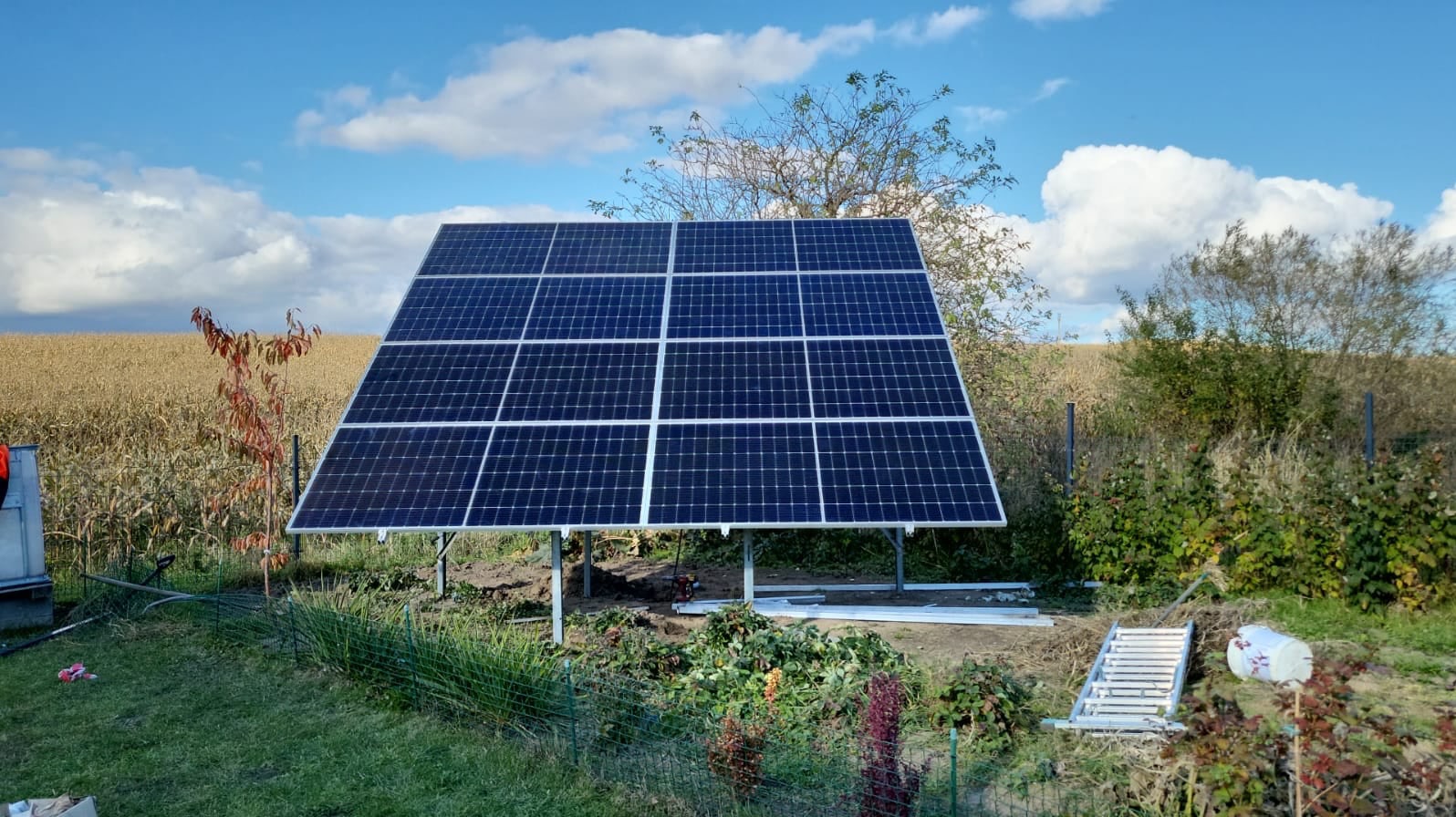 Panel fotowoltaiczny na metalowej konstrukcji, widoczny na tle pola uprawnego i pochmurnego nieba, częściowo ogrodzony niskim płotkiem, z elementami montażowymi w pobliżu.