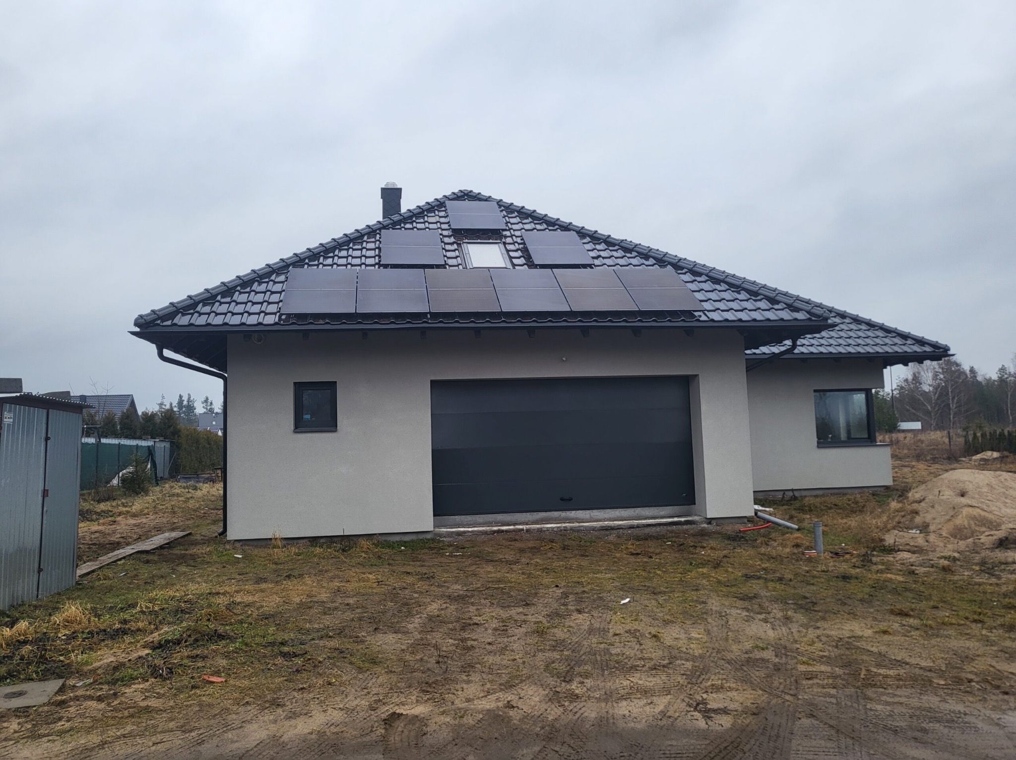 Dom jednorodzinny z ciemną dachówką i panelami słonecznymi na dachu, widok z przodu w pochmurny dzień, teren wokół domu częściowo zagospodarowany.