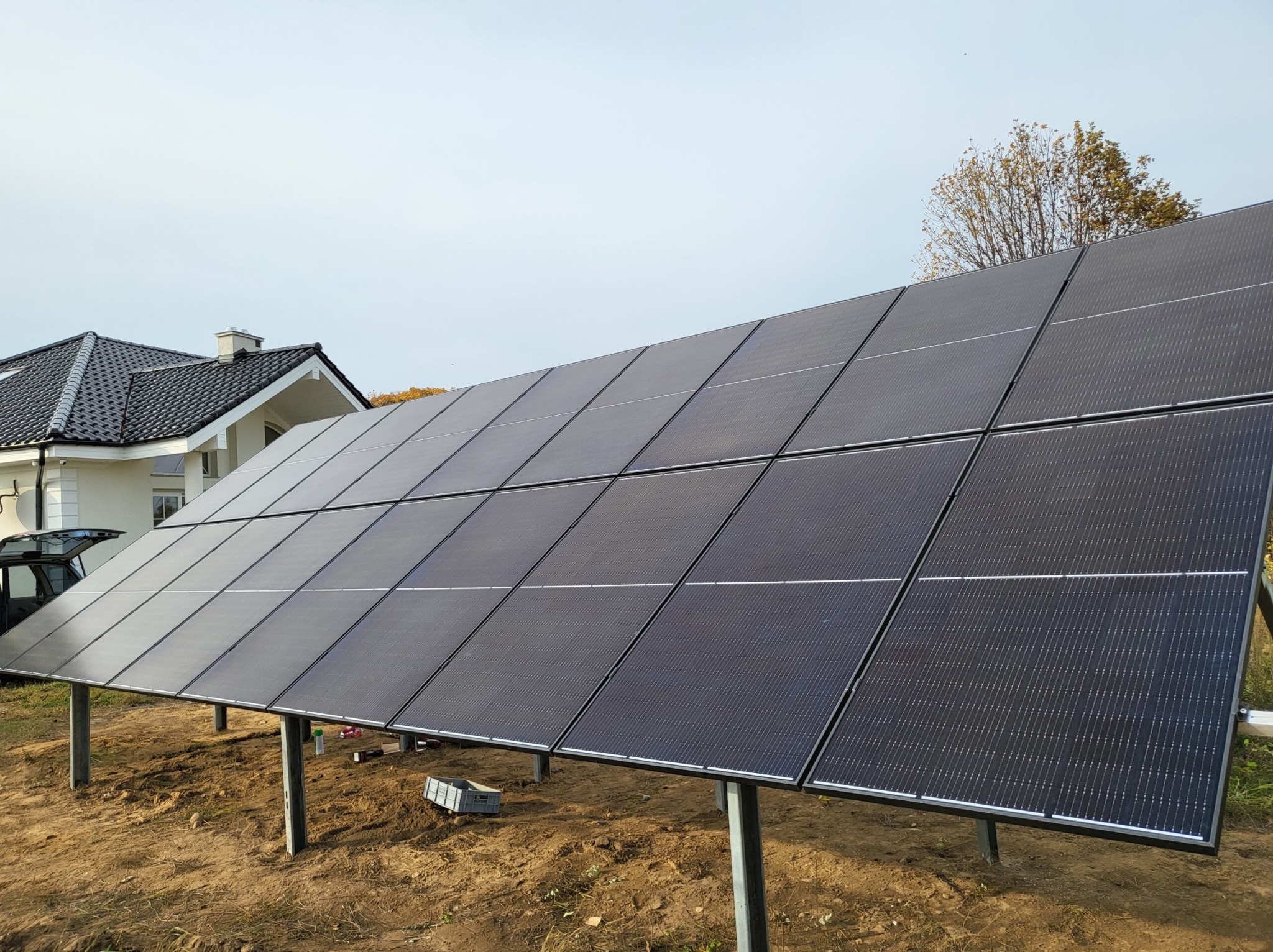 Rząd paneli słonecznych na metalowych słupkach, z domem z szarą dachówką w tle, skrzynka narzędziowa na ziemi sugeruje trwający montaż.