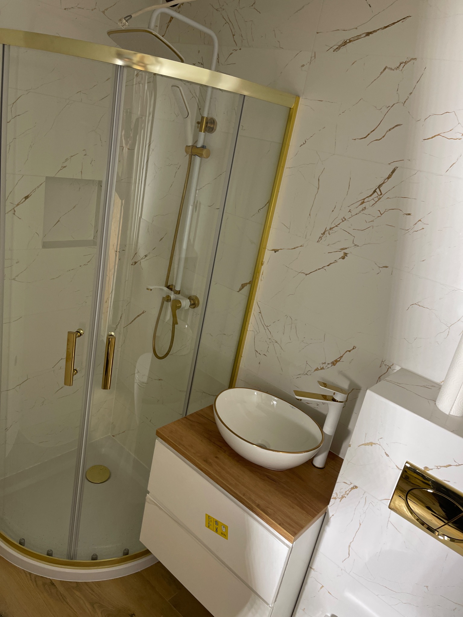 Nowoczesna łazienka z kabiną prysznicową ze złotymi ramami, umywalką nablatową na drewnianym blacie i złotymi akcentami na białych marmurowych płytkach ściennych.