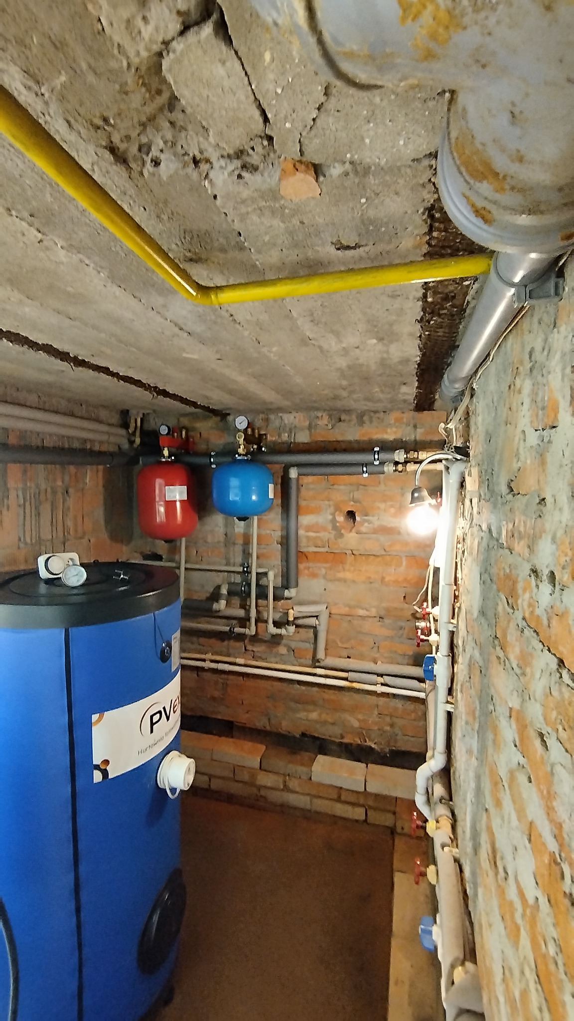 Instalacja w piwnicy z nieotynkowaną cegłą, widoczny zbiornik w niebieskiej obudowie z naklejoną etykietą, czerwony i niebieski zbiornik wyrównawczy, rury w różnych kolorach, surowy betonowy sufit...