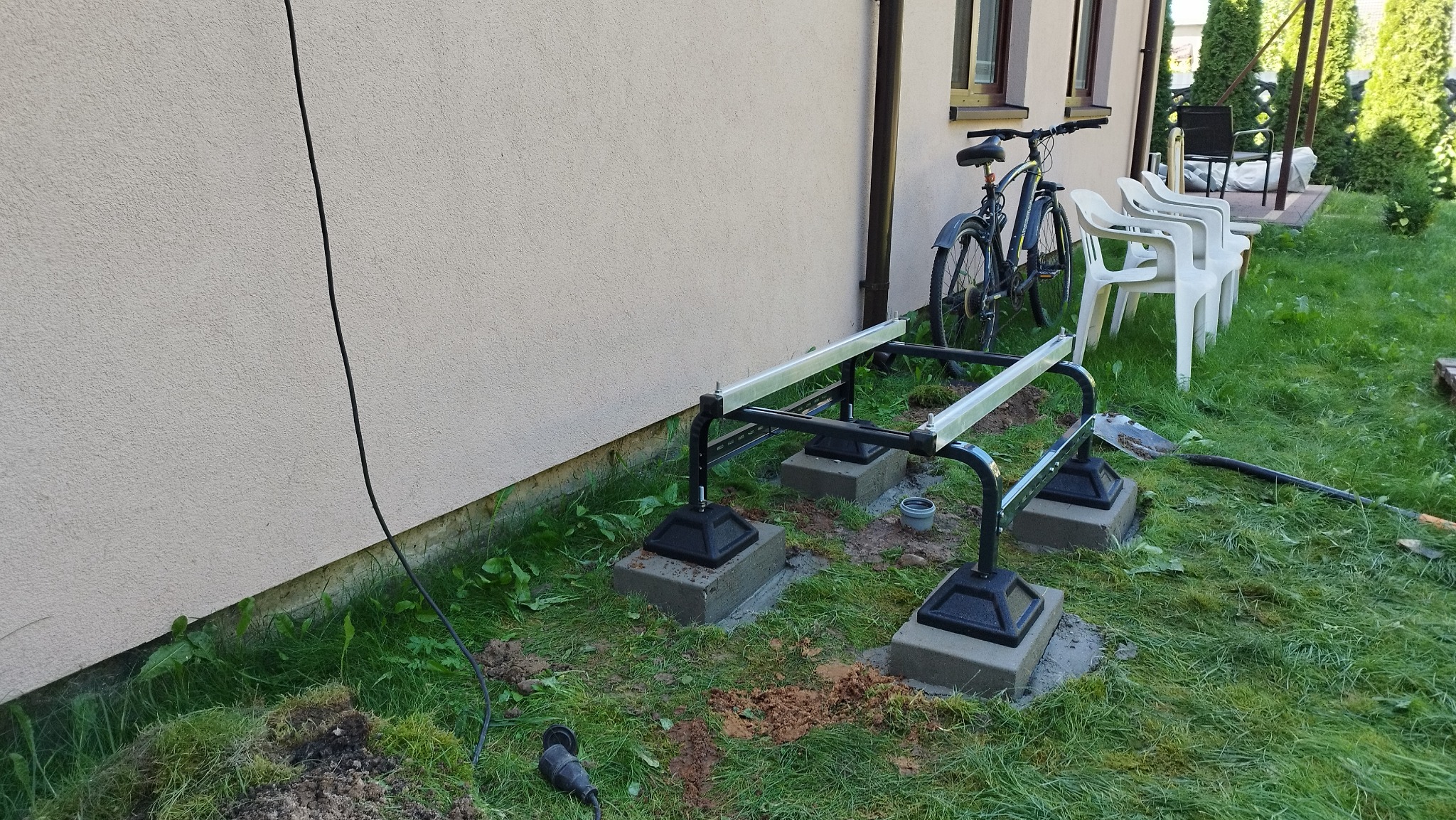 Przygotowanie fundamentów pod konstrukcję tarasu przy ścianie domu, widoczne betonowe podstawy z czarnymi wspornikami i aluminiowymi profilami, na trawniku leży łopata, w tle rower i białe krzesła.