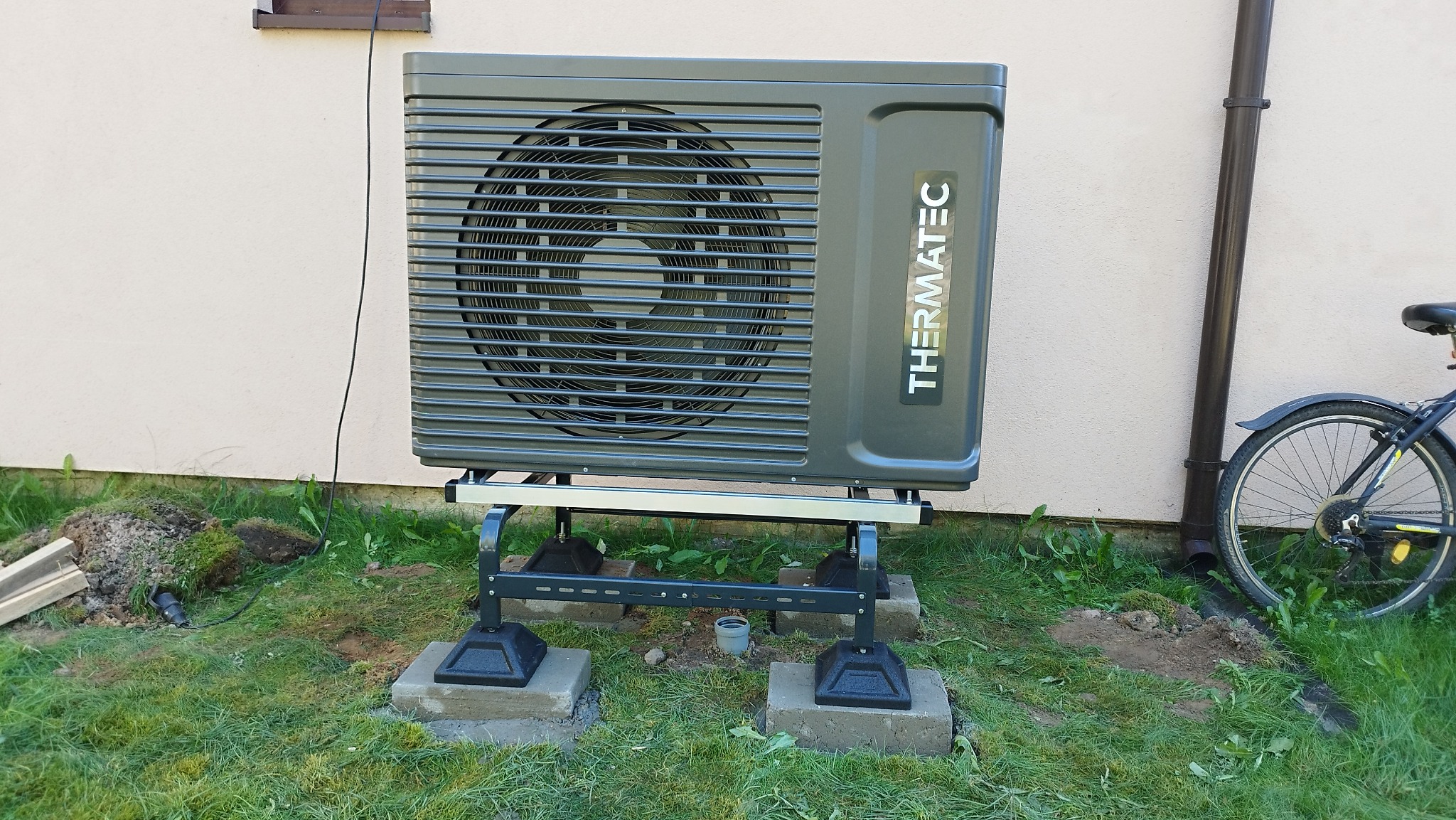 Szara jednostka zewnętrzna z logo THERMATEC, umieszczona na metalowej konstrukcji na betonowych podstawach, obok ściany budynku i rury spustowej, z fragmentem roweru w tle.
