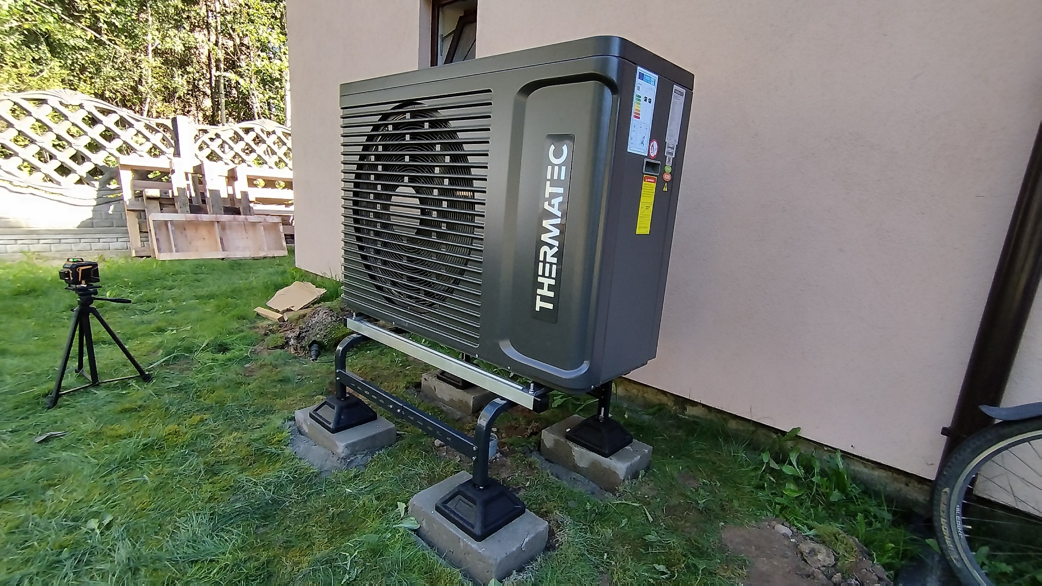 Szara jednostka zewnętrzna z logo THERMATEC, zamontowana na metalowej konstrukcji z betonowymi podstawami na trawniku, obok ściany budynku. Widoczny niwelator laserowy na statywie.