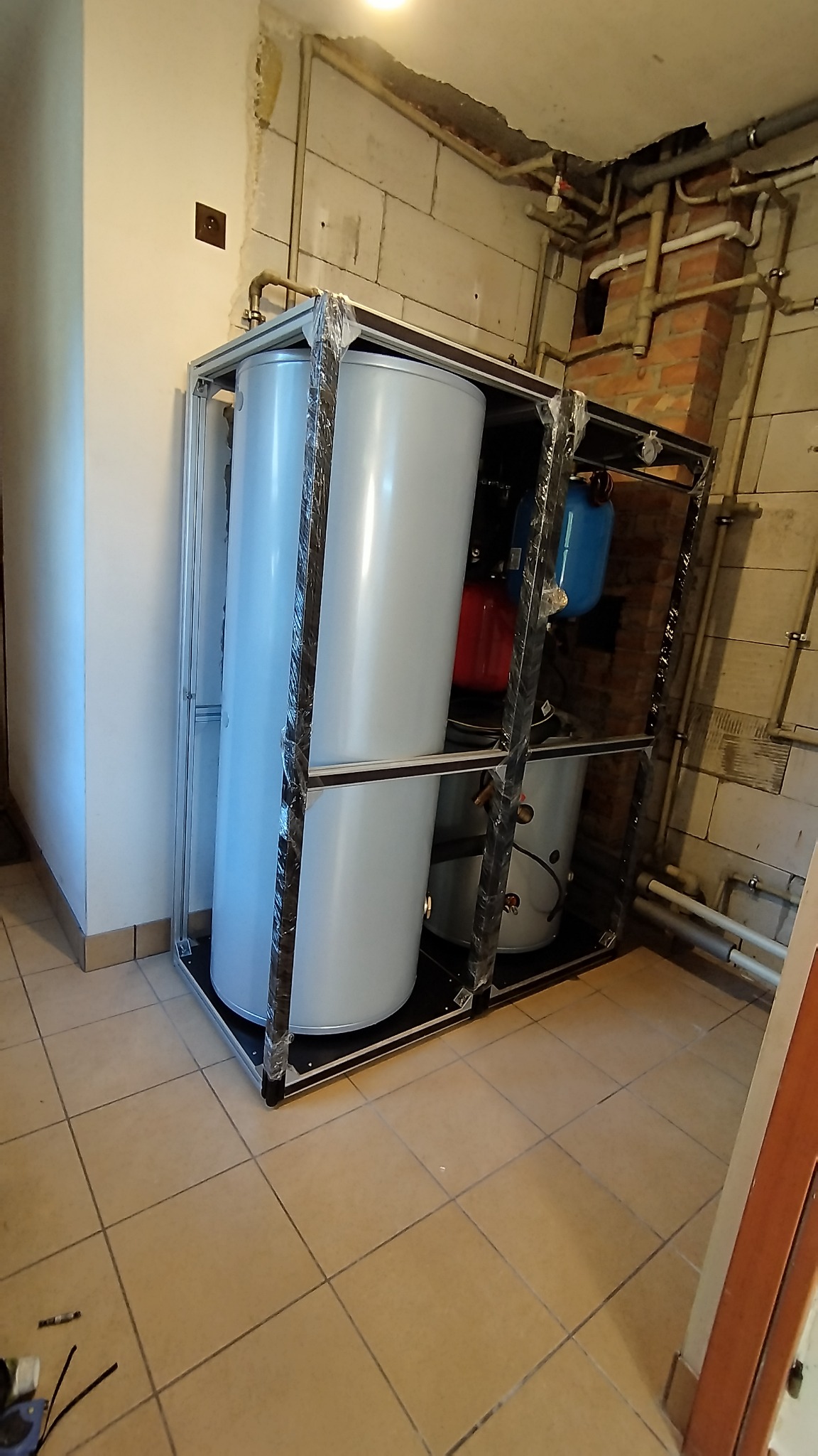 Kompaktowa instalacja pompy ciepła z dwoma zbiornikami buforowymi, w metalowej ramie, z widocznymi elementami hydraulicznymi w tle, na tle surowej ściany z instalacjami.