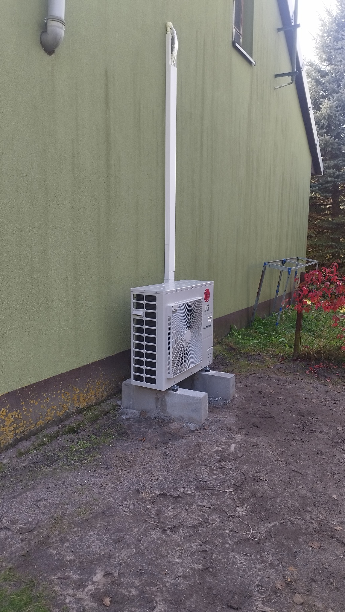 Zewnętrzna jednostka pompy ciepła LG Therma V, zamontowana na betonowych podstawach przy ścianie budynku o oliwkowym kolorze, z widoczną rurą odprowadzającą kondensat.