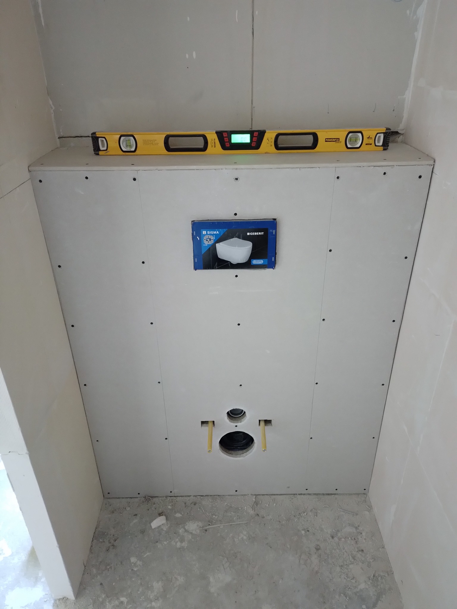 Stelaż podtynkowy Geberit zabudowany płytą gipsowo-kartonową, widoczny poziomica Powerfix, otwory na przyłącza i opakowanie po ceramice.