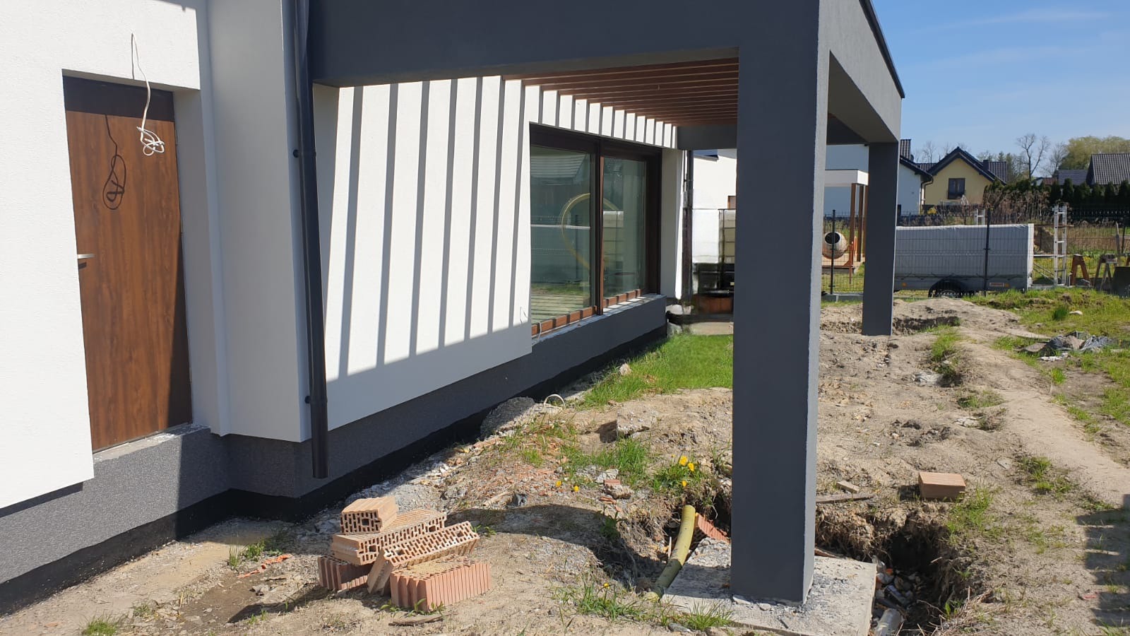 Nowoczesny dom jednorodzinny w trakcie wykończenia elewacji, widoczne cegły, świeżo pomalowane ściany w kolorze białym i szarym oraz drewniane elementy dekoracyjne.