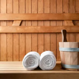 Wnętrze sauny z drewnianą boazerią, dwoma zwiniętymi białymi ręcznikami i drewnianym wiadrem z nabierką i chochlą.