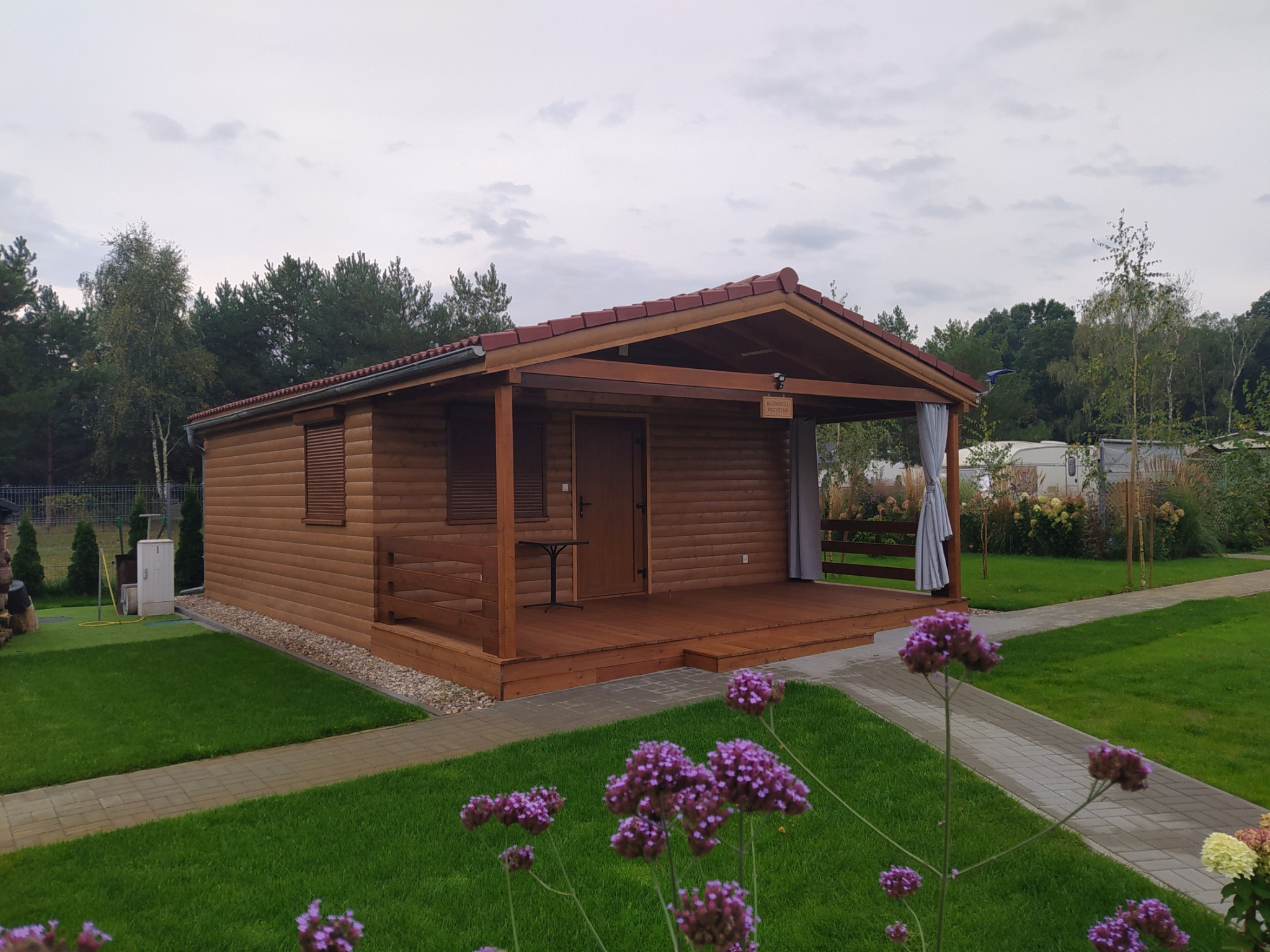 Drewniany domek letniskowy z zadaszonym tarasem, wykończony w naturalnym brązie, z czerwoną dachówką, otoczony zielonym trawnikiem i rabatami kwiatowymi. Na tarasie mały stolik, a z boku zwisająca...