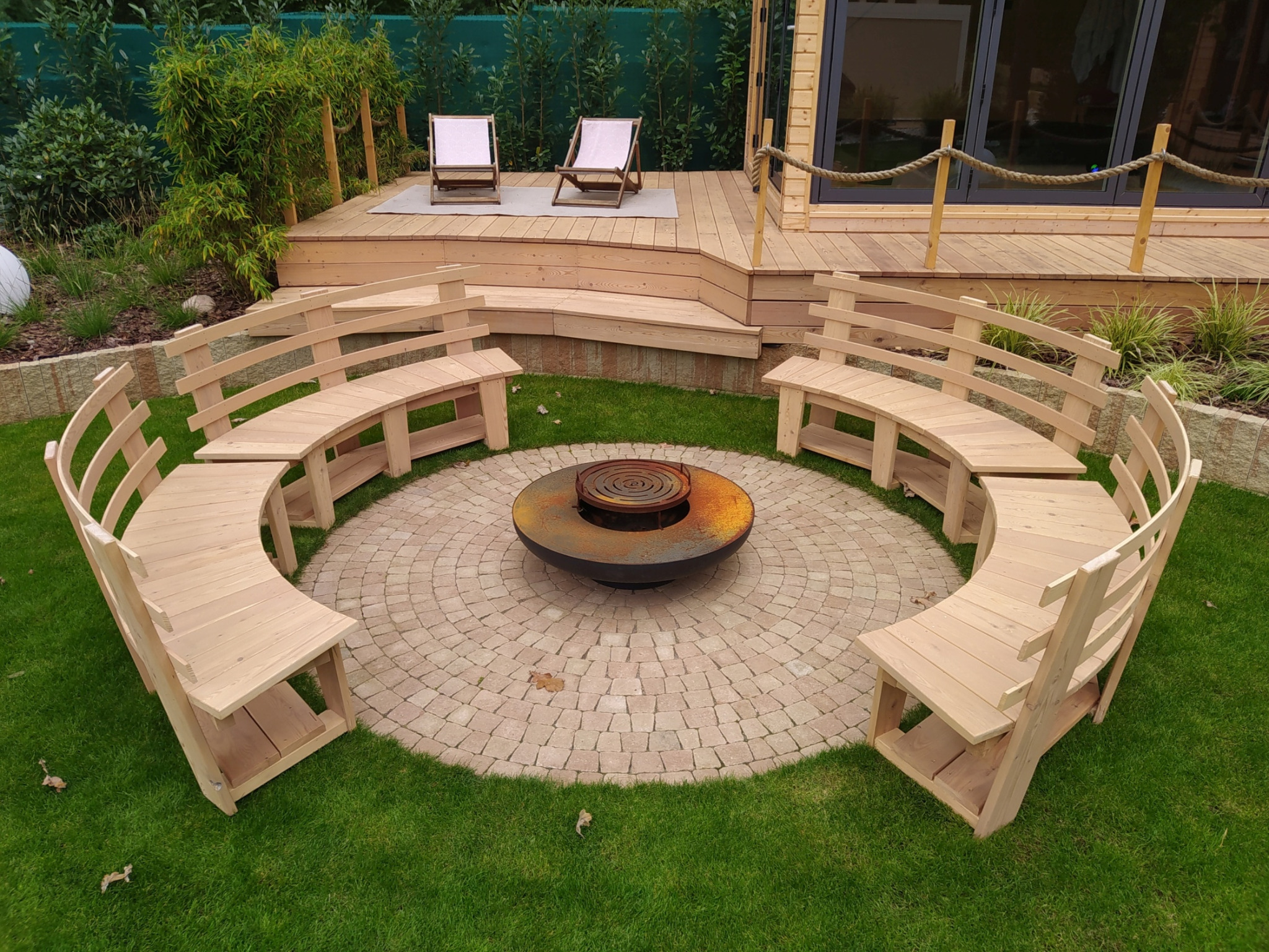 Ogród z okrągłym paleniskiem otoczonym drewnianymi, łukowymi ławkami, w tle drewniany taras z dwoma leżakami i domkiem z dużymi oknami, zielona roślinność dookoła.