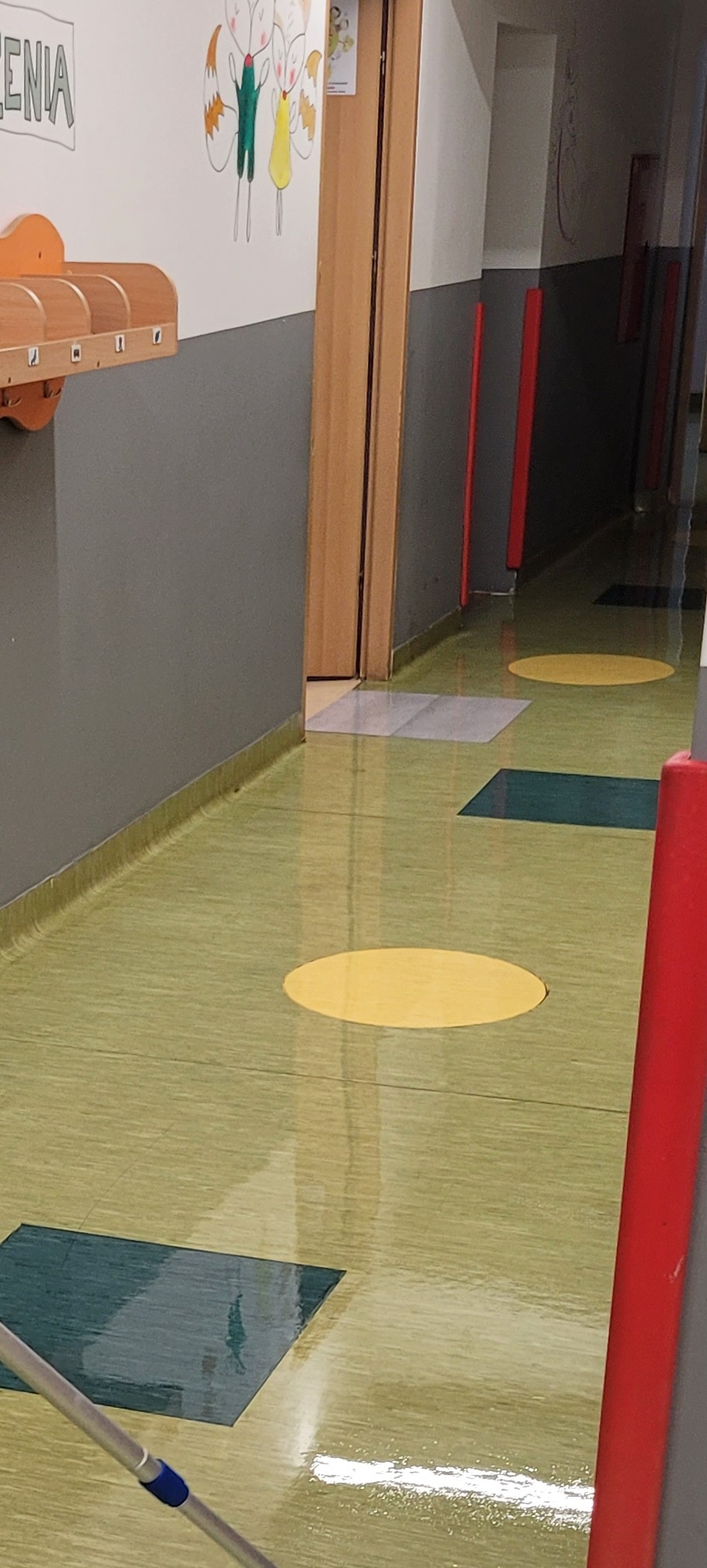 Mop na mokrej podłodze w korytarzu z żółtymi okręgami i zielonymi kwadratami, ślady wody na podłodze, widoczne odbicia światła.