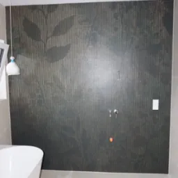 Ściana łazienki wyłożona ciemnymi płytkami z motywem liści na tle złotych, pionowych pasków, widoczna bateria prysznicowa i biała wanna wolnostojąca, częściowo zakryta folią ochronną lampa sufitowa.