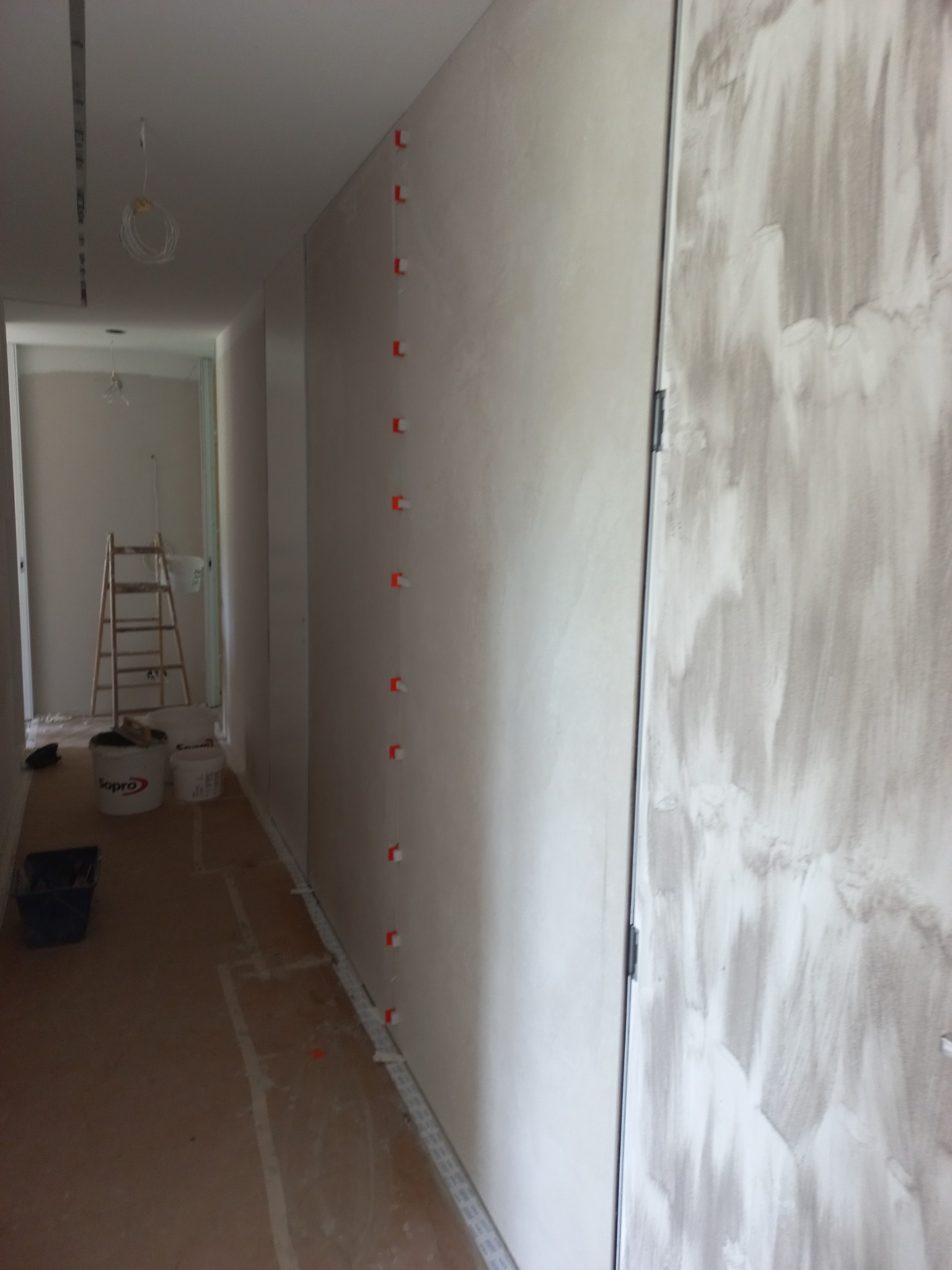 Wnętrze w trakcie prac wykończeniowych: ściana z pionowym rzędem klipsów poziomujących płytki, widoczne wiadra z zaprawą i drabina w głębi pomieszczenia.