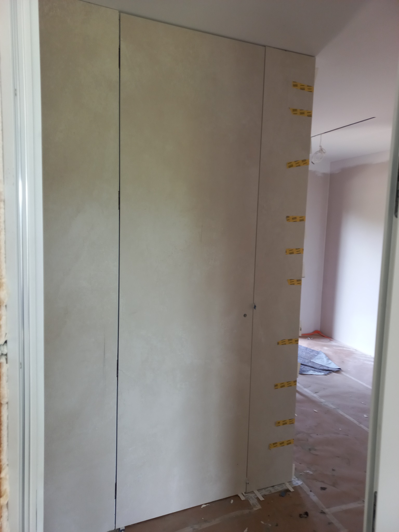 Ściana z płyt gipsowo-kartonowych w trakcie montażu, widoczne żółte taśmy mocujące na krawędziach, przygotowana do dalszych prac wykończeniowych w jasnym wnętrzu.