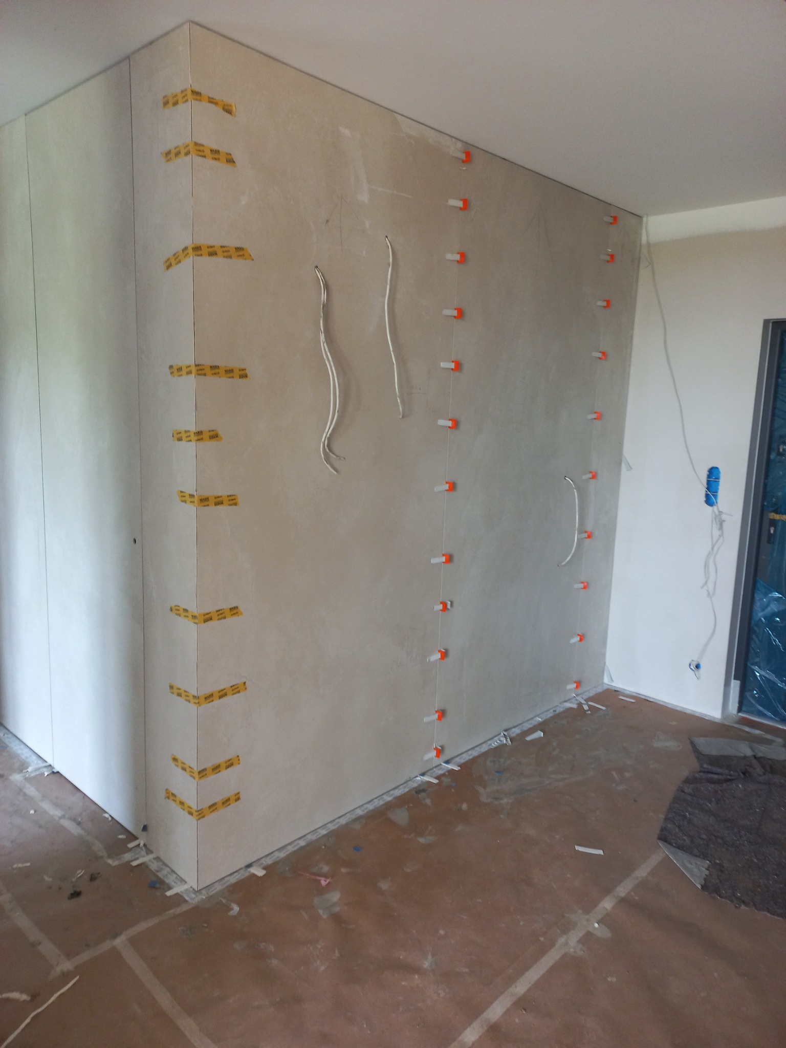 Ściana w trakcie układania płytek, widoczne poziomowanie za pomocą klipsów, taśmy maskujące na rogach, wystające przewody elektryczne.