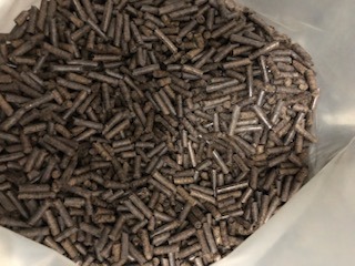 Wsypany do przezroczystego worka pellet drzewny o nieregularnym kształcie i ciemnobrązowej barwie, widoczne drobne fragmenty i pył.