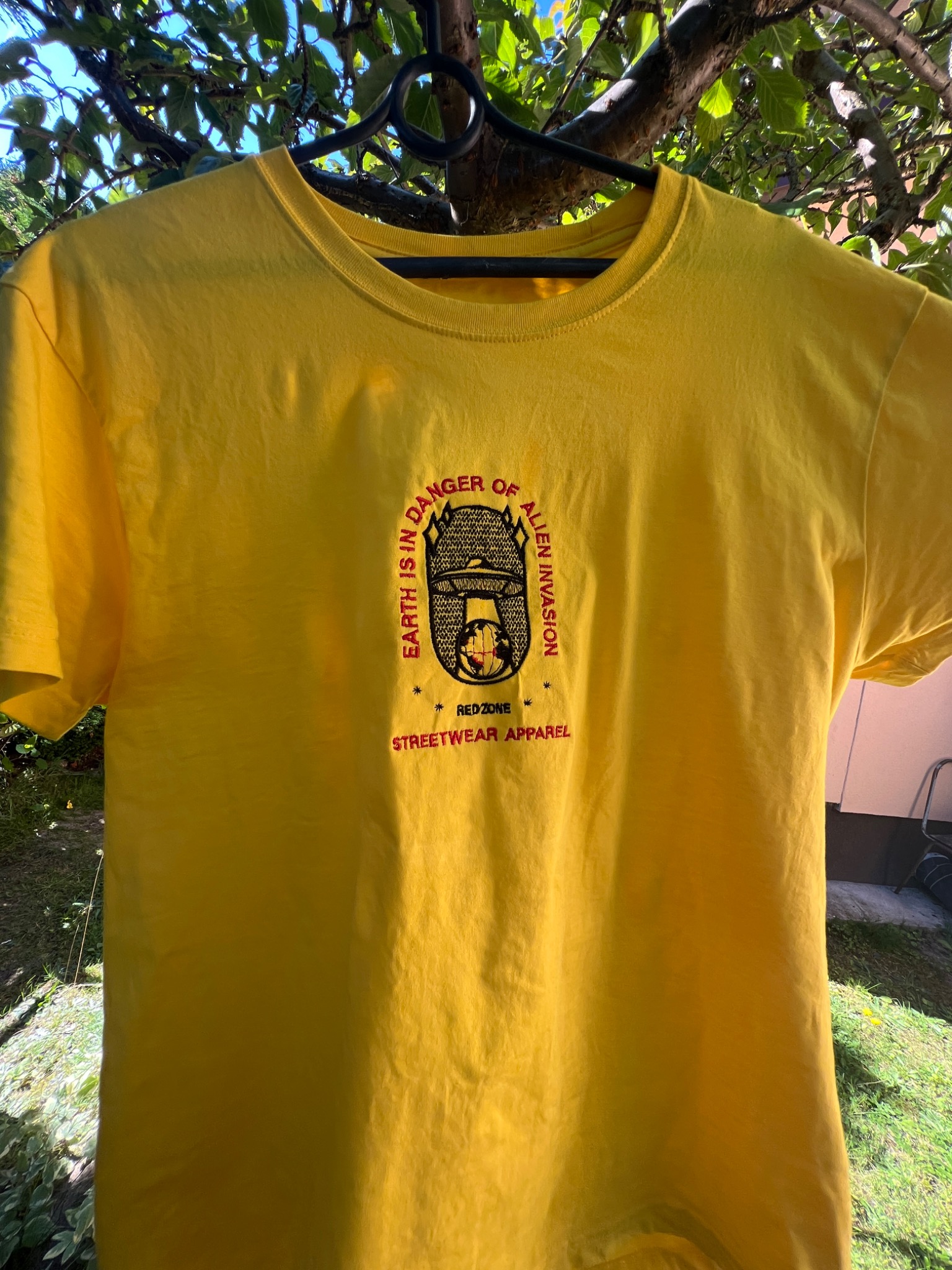 Żółta koszulka z haftowanym motywem UFO i napisem 'Earth is in danger of alien invasion, Red Zone Streetwear Apparel', zawieszona na wieszaku na tle zieleni.