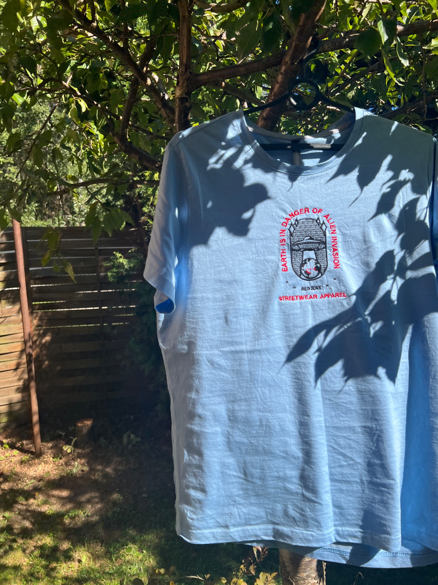 Jasnoniebieska koszulka z haftowanym motywem UFO i napisem 'Earth is in danger of alien invasion' oraz 'Red Zone Streetwear Apparel', wisząca na wieszaku na tle zieleni i drewnianego płotu.