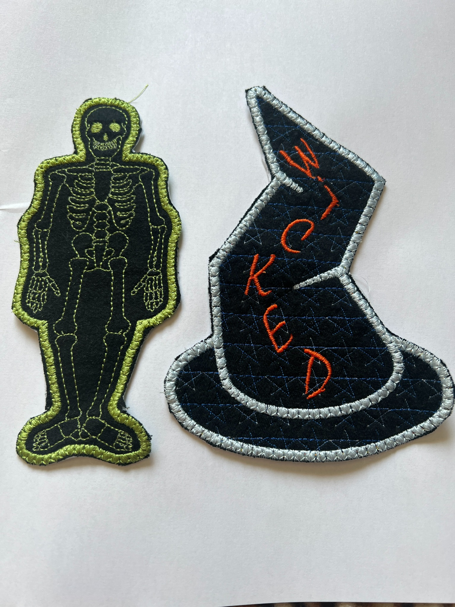 Dwa naszywane, filcowe motywy: czarny szkielet z neonowo-zielonym obrysem i czarny kapelusz czarownicy z napisem 'WICKED' haftowanym pomarańczową nicią, oba na białym tle.