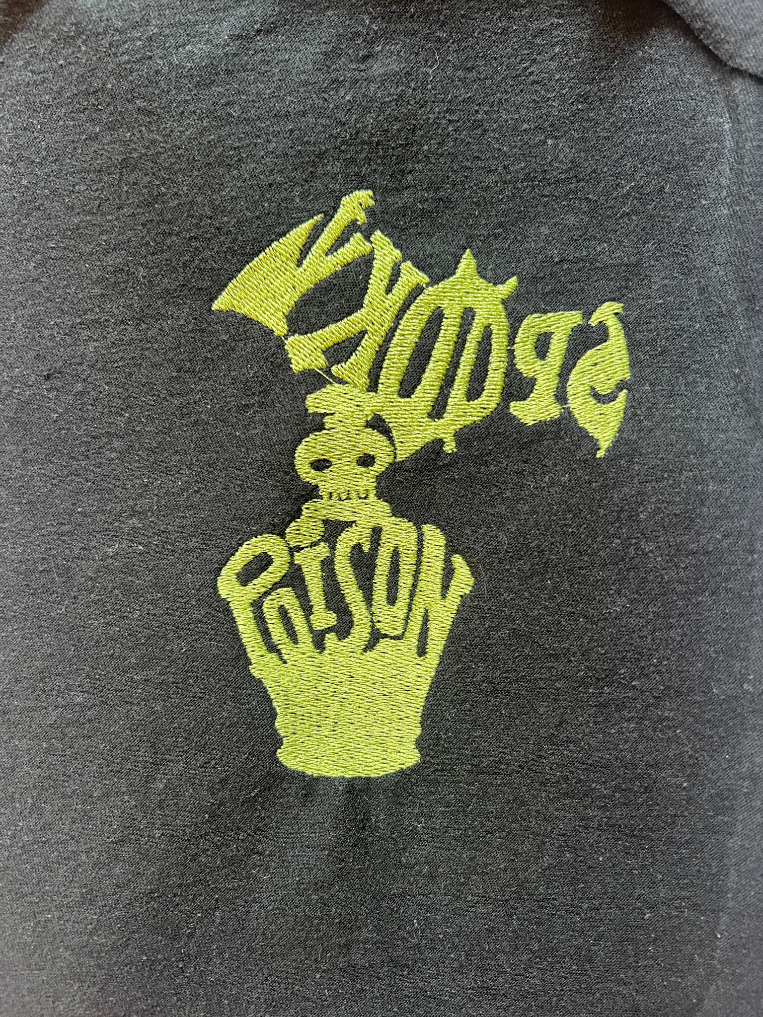 Czarna koszulka z haftowanym, jaskrawozielonym napisem 'EXODUS PRISON' ułożonym w kształt czaszki i dłoni.