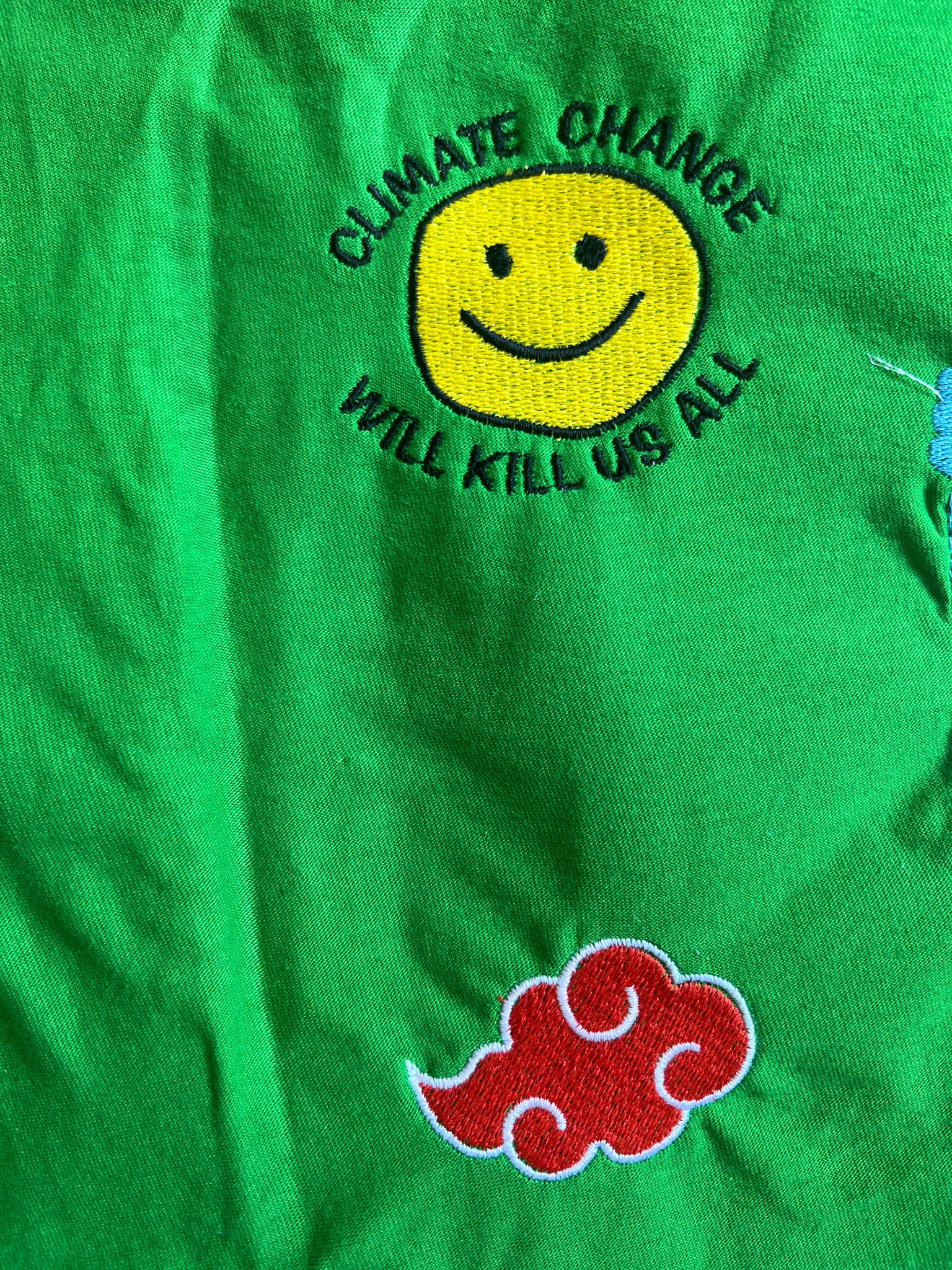 Jasnozielona tkanina z wyhaftowanym żółtym uśmiechem otoczonym napisem 'CLIMATE CHANGE WILL KILL US ALL' oraz czerwoną chmurą z białym obrysem, nawiązującą do motywu z anime.