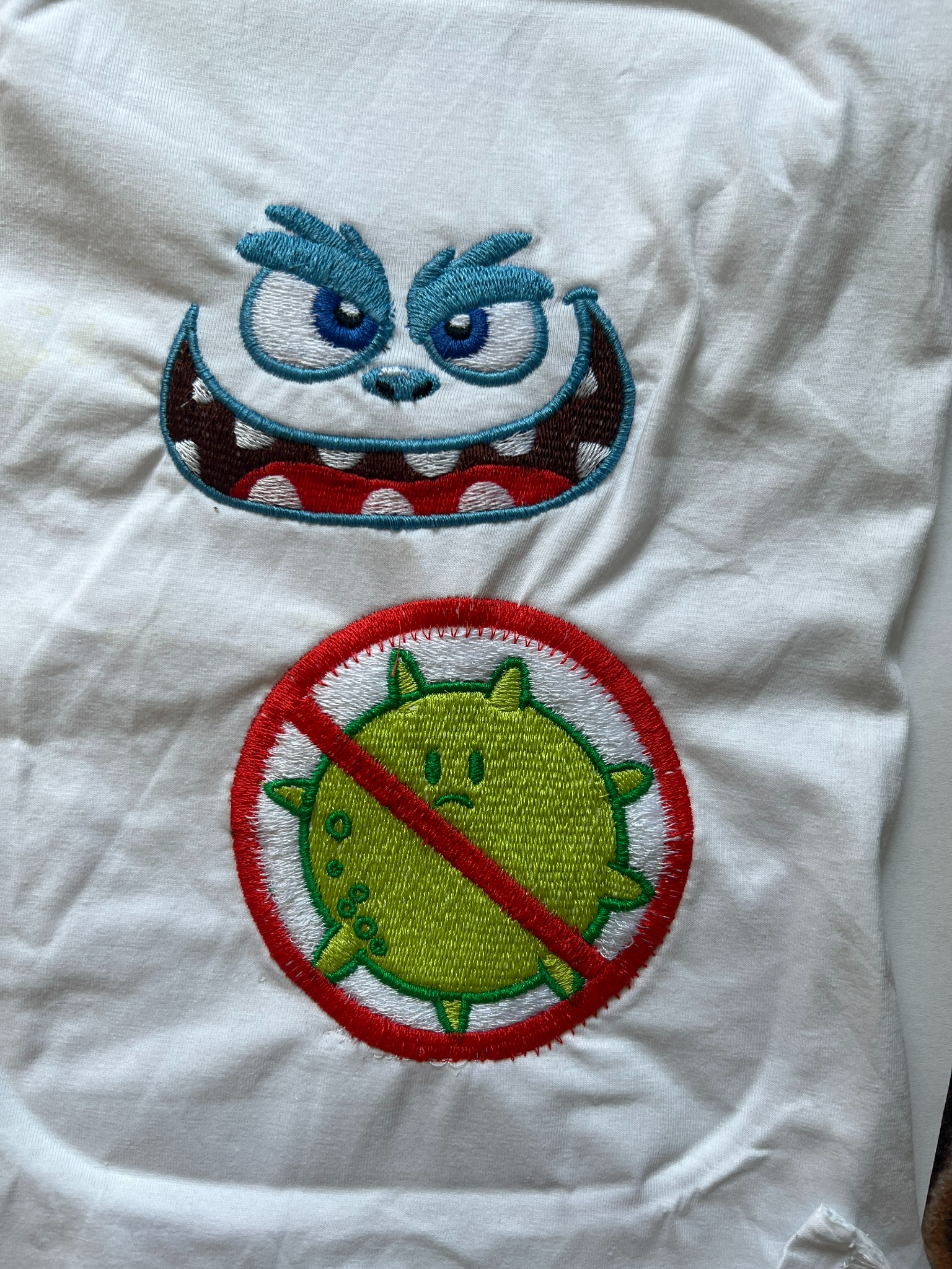 Biała koszulka z haftowanym, kreskówkowym potworem z niebieskimi oczami i zielonym wirusem przekreślonym czerwoną linią.