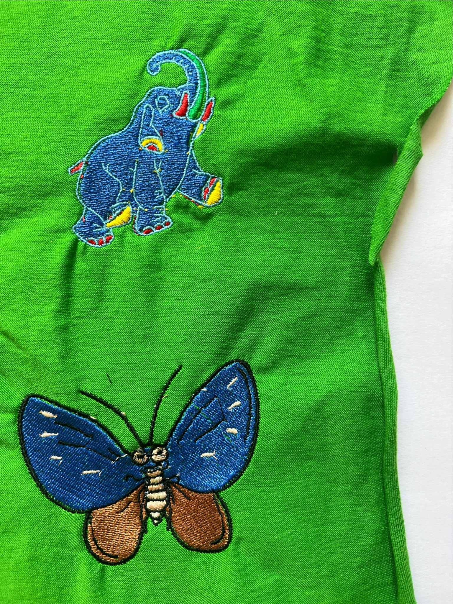 Jasnozielona koszulka z haftowanym niebieskim słoniem z podniesioną trąbą i brązowo-niebieskim motylem.