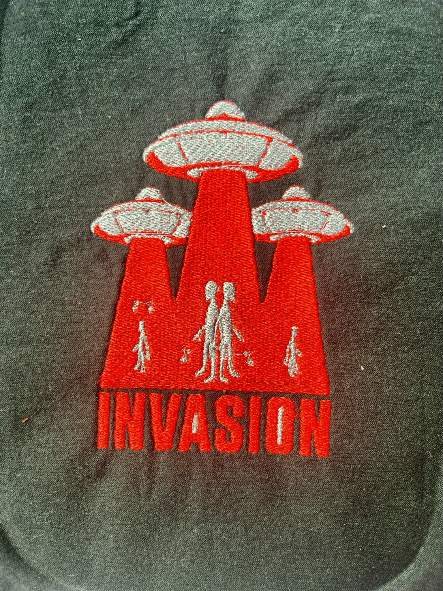 Czarne ubranie z czerwono-szarym haftem przedstawiającym trzy latające spodki z promieniami i sylwetkami kosmitów oraz napisem 'INVASION'.