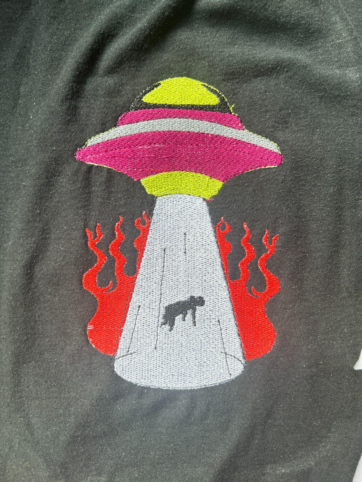 Czarna koszulka z haftowanym kolorowym motywem UFO z wiązką światła i sylwetką zwierzęcia, otoczonego czerwonymi płomieniami.