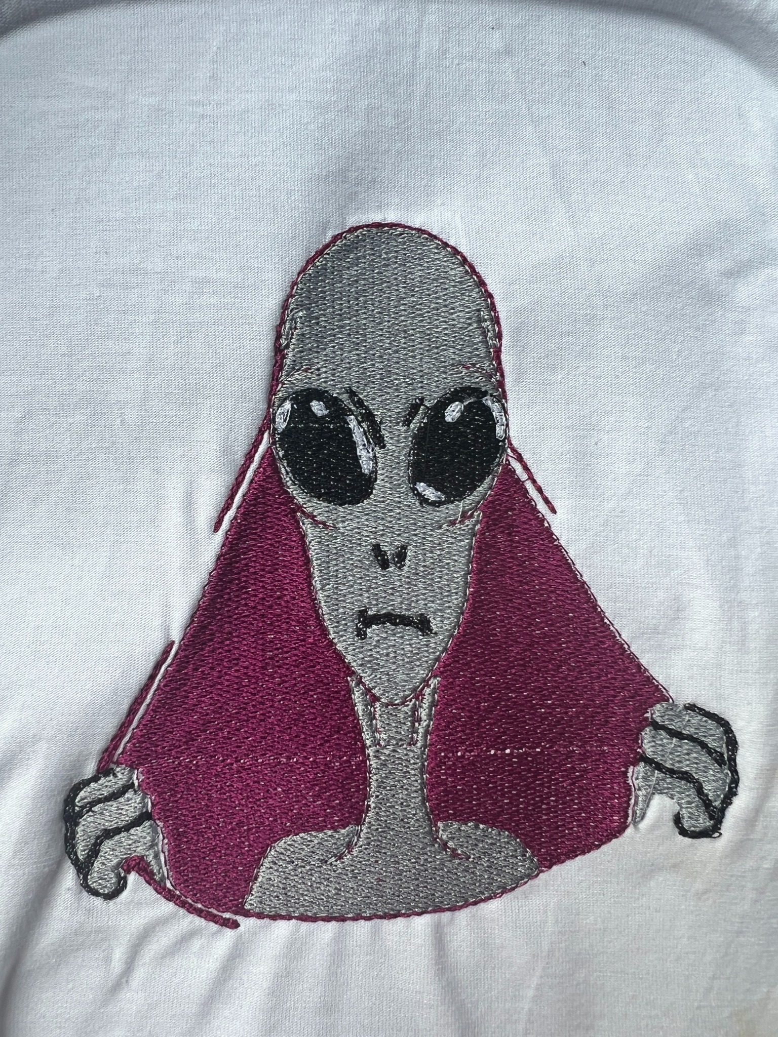Biała tkanina z wyhaftowanym motywem szaro-fioletowego kosmity z dużymi czarnymi oczami i ponurym wyrazem twarzy.