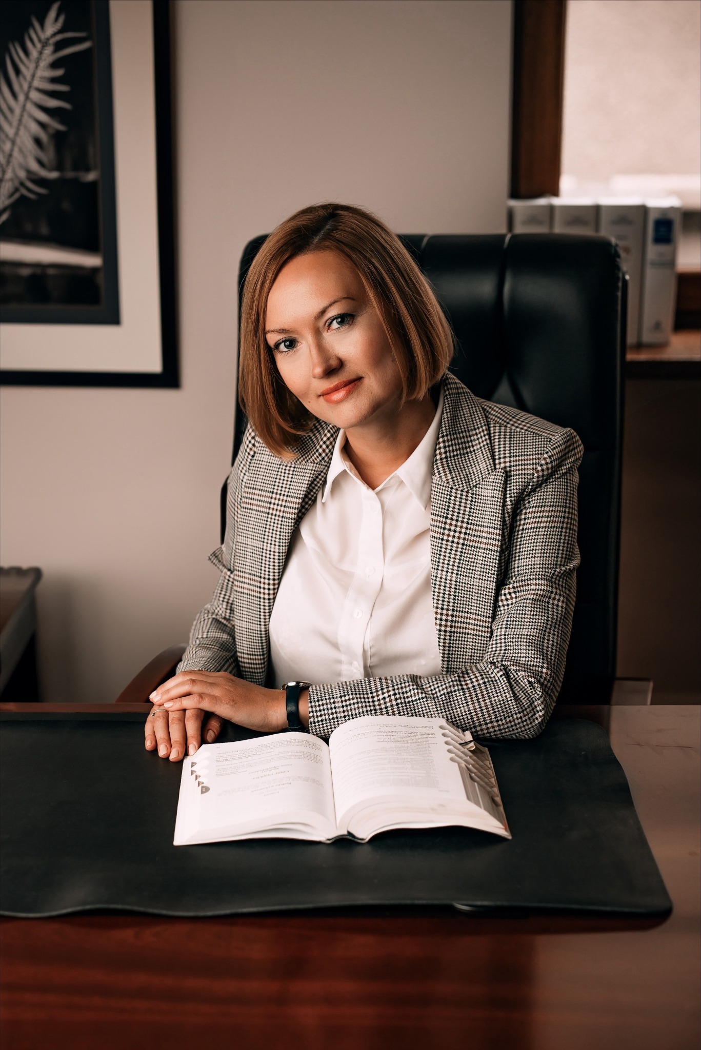 Kobieta w kraciastej marynarce siedzi za biurkiem z otwartą książką, na tle biura z obrazem i regałem z segregatorami.